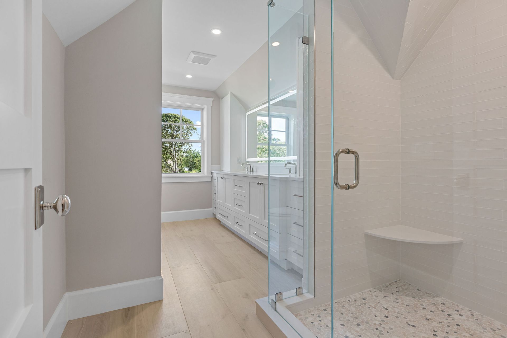 A modern bathroom featuring a white vanity with a lighted mirror, a glass-enclosed shower, and light wood flooring.