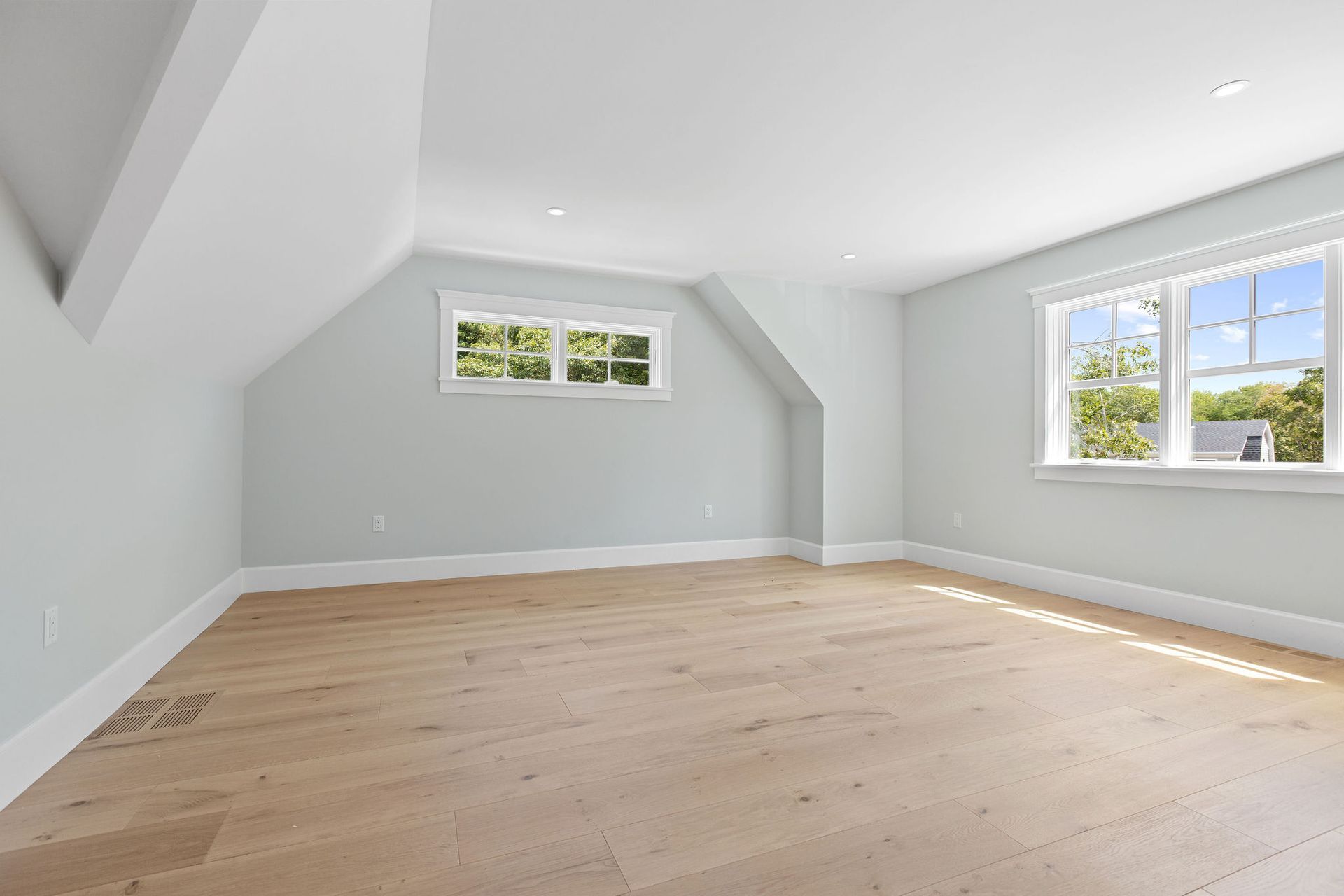 An empty attic room with light wood floors, slanted white walls, light gray accent walls, and two windows.