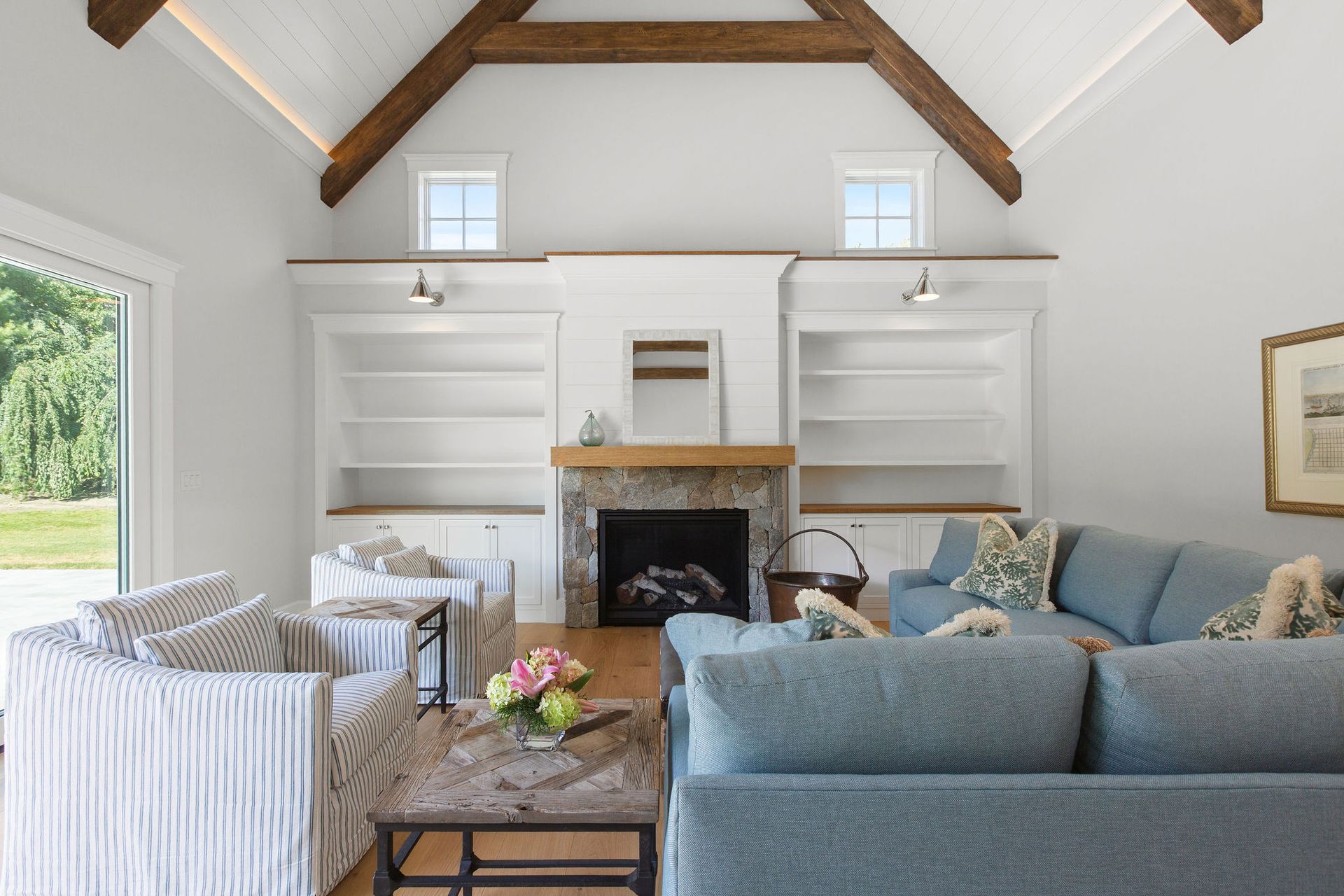 A cozy living room with a vaulted ceiling, exposed beams, a stone fireplace, built-in shelves, and blue and white furniture.