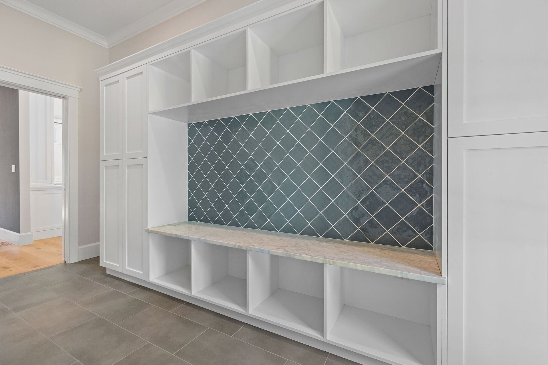 White mudroom built-in storage with a bench, open cubbies, and a dark, diamond-patterned tiled backsplash.