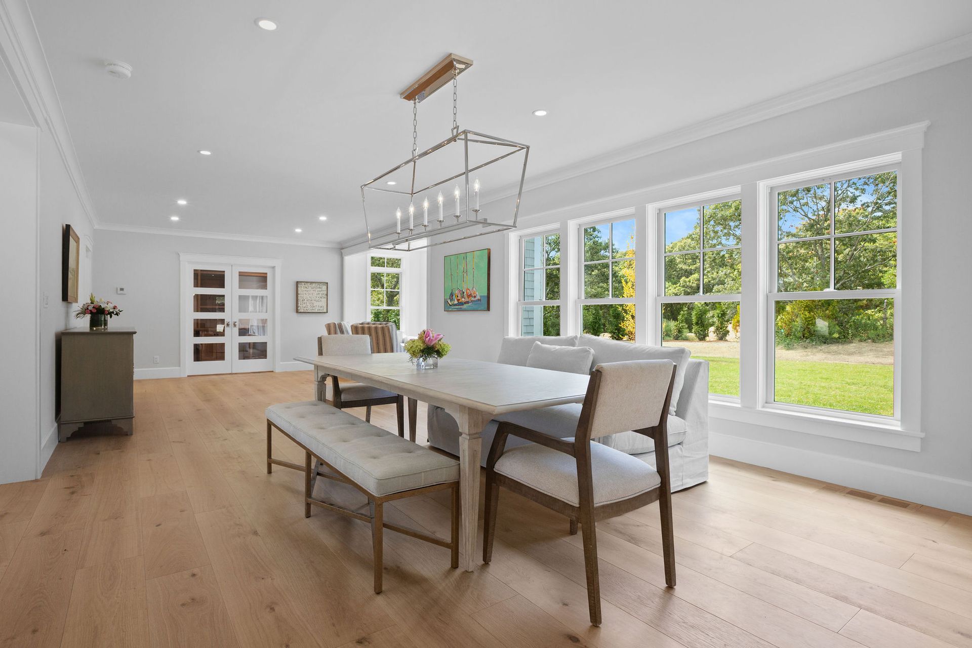 A bright dining room with light wood floors, a wooden table with a bench and chairs, and large windows looking outside.