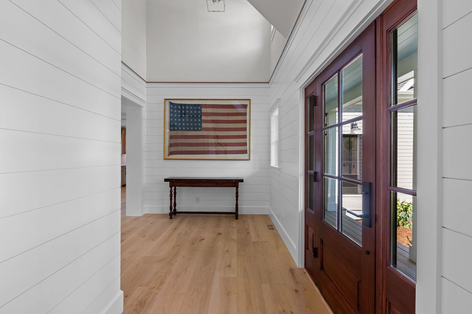 A bright entryway with white shiplap walls, light wood floors, a framed US flag on the wall, and a wood-paned glass door.