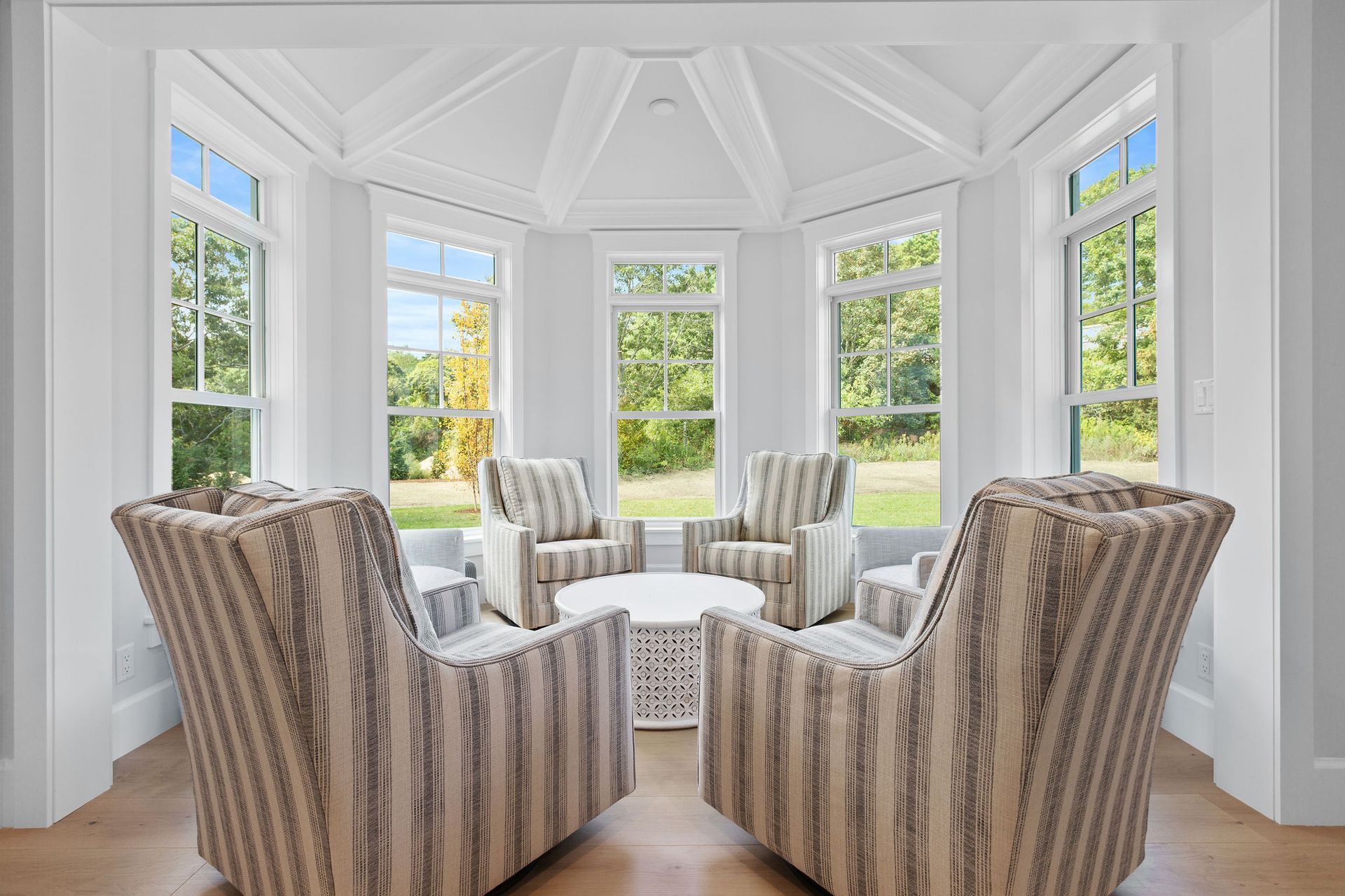 A bright, white sunroom featuring four patterned armchairs arranged around a round white table in front of large windows.
