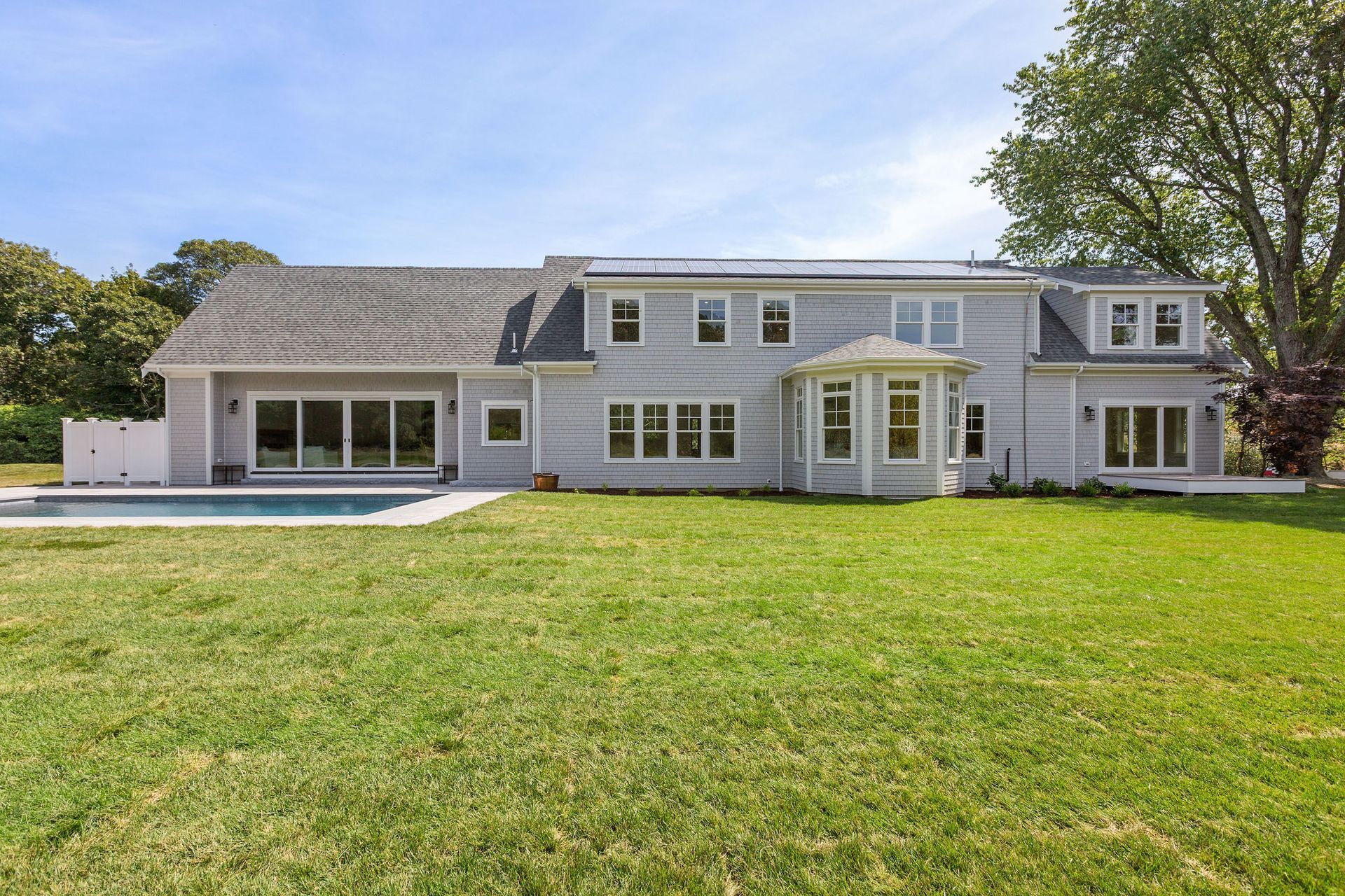 A two-story light gray house with a large backyard and swimming pool under a bright blue sky.