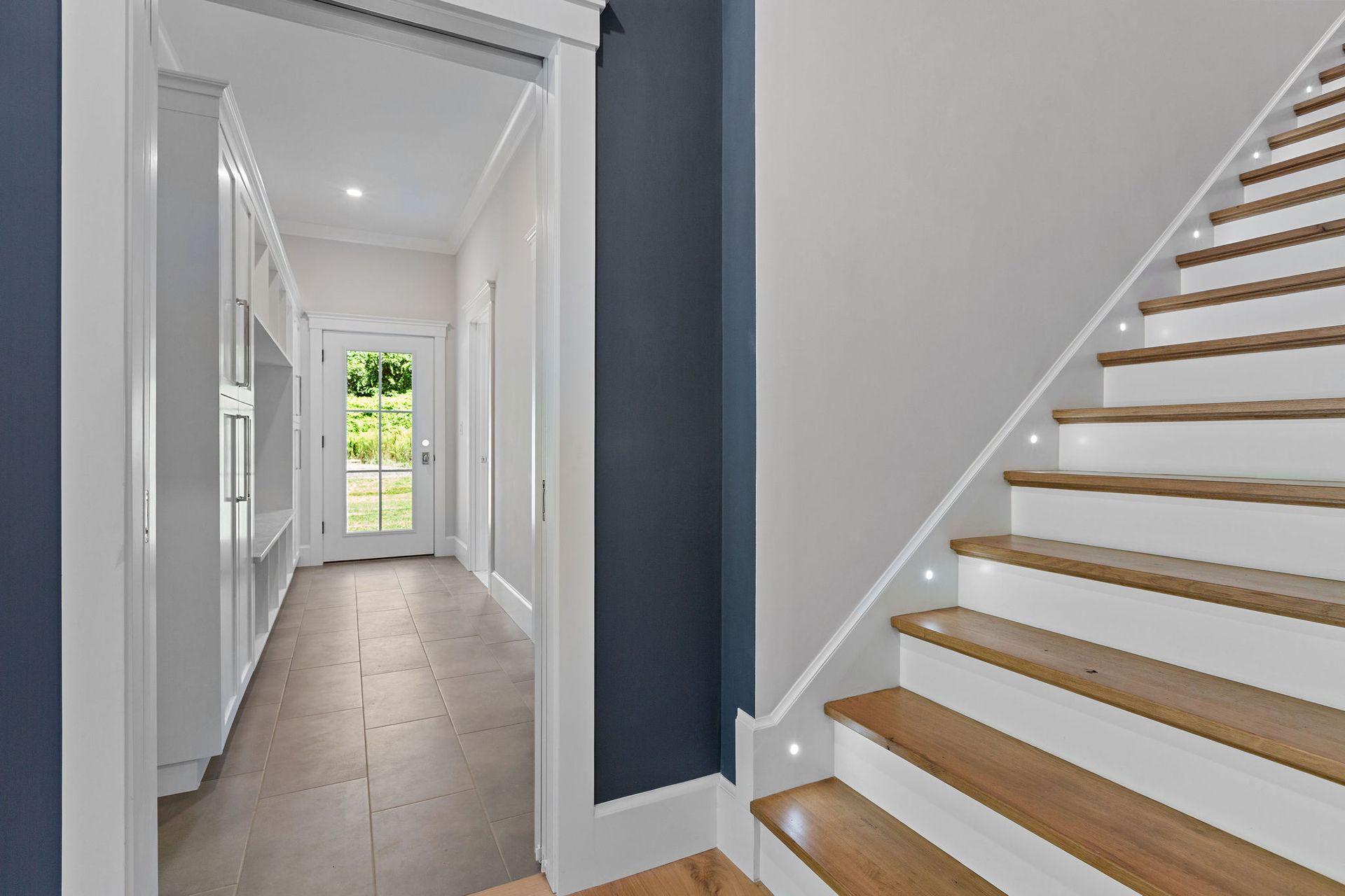 A view of a modern home entryway with wooden stairs, white walls, navy blue accents, and a hallway leading to a front door.