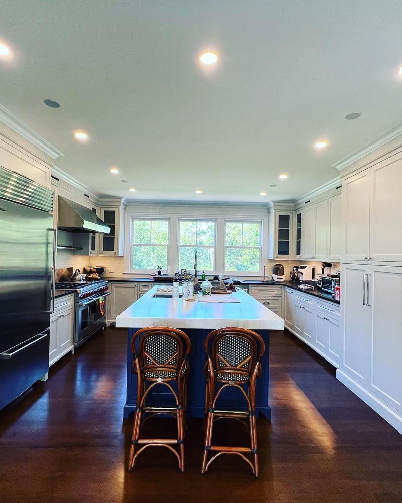 A bright kitchen with white cabinets, dark wood floors, a blue island with two stools, and stainless steel appliances.