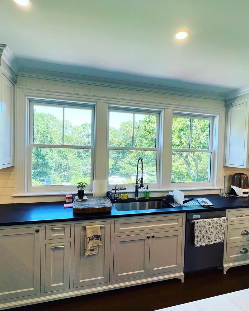 A kitchen sink area with a large three-panel window, light-colored cabinets, and dark countertops.