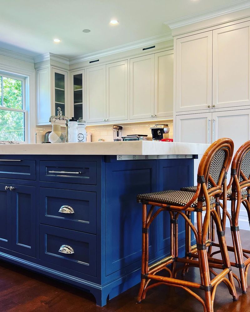 A dark blue kitchen island with a light countertop sits in front of white cabinets and two woven rattan bar stools.