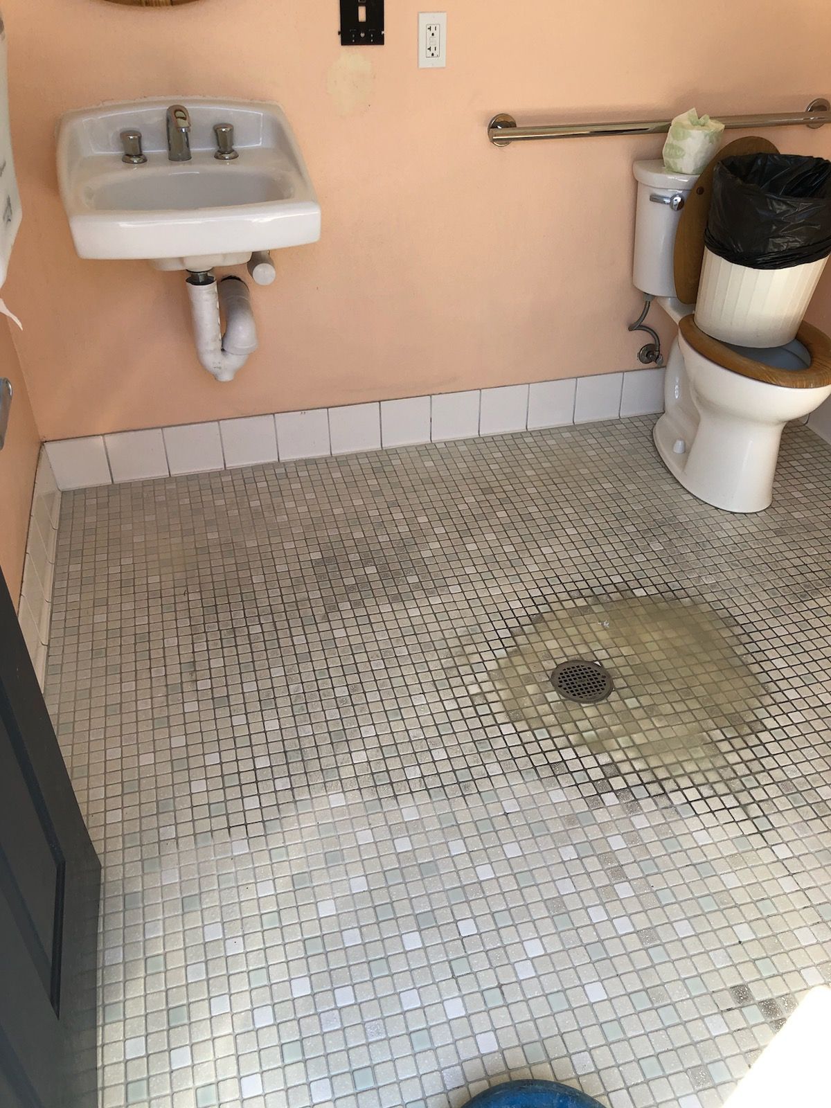 Public restroom with wall sink, toilet, and tiled floor drain, showing visible floor stains.