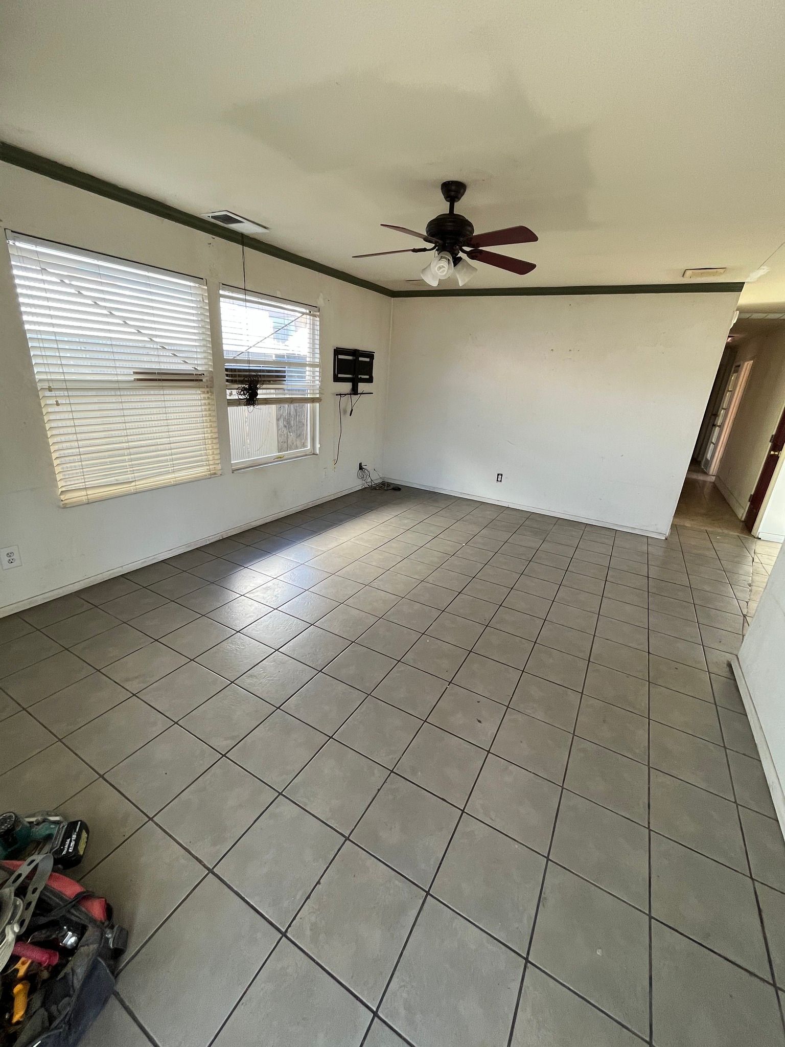 Empty tiled living room with white walls, ceiling fan, and windows with blinds.