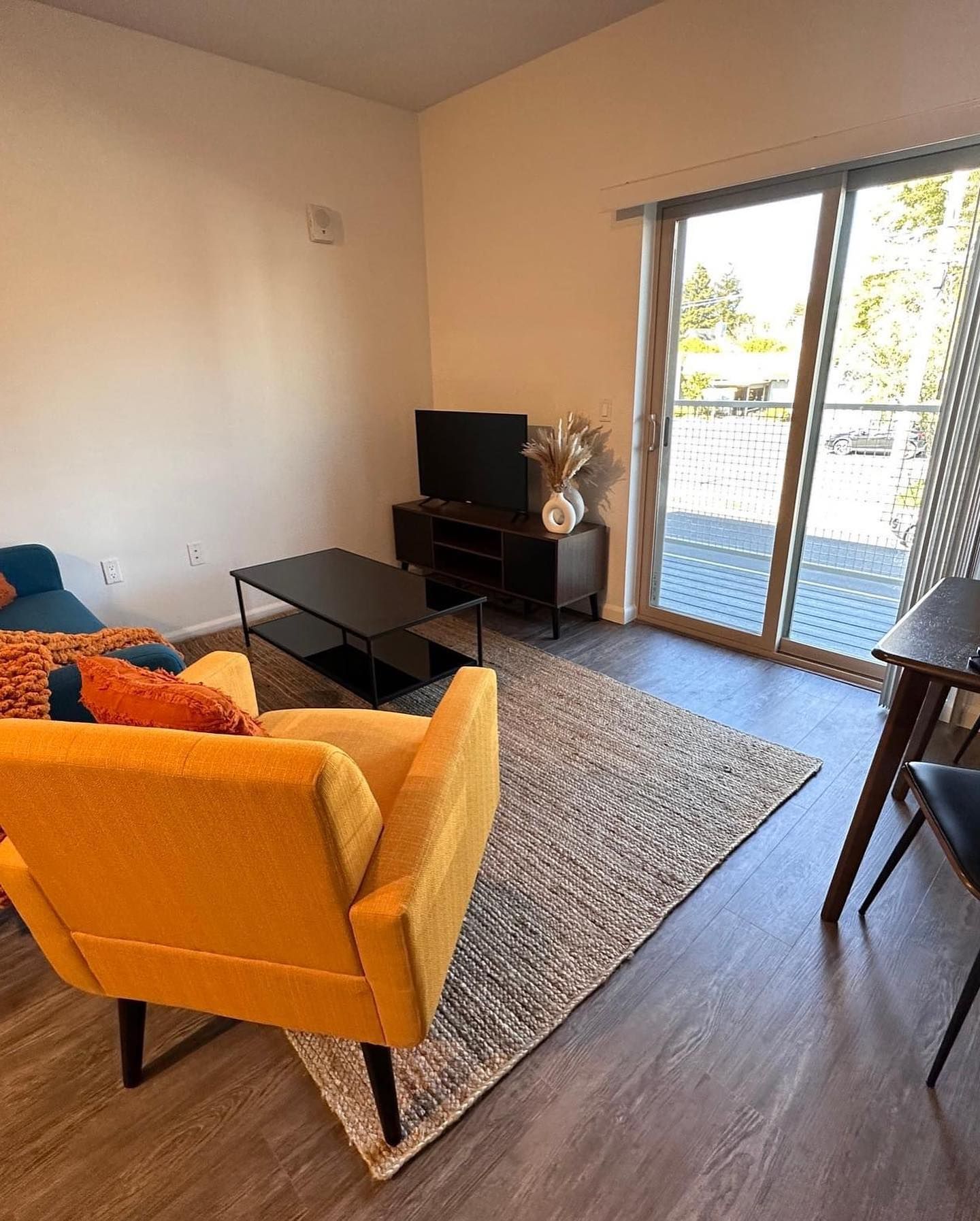 Modern living room with yellow sofa, black TV stand, coffee table, rug, and balcony doors open to outside.