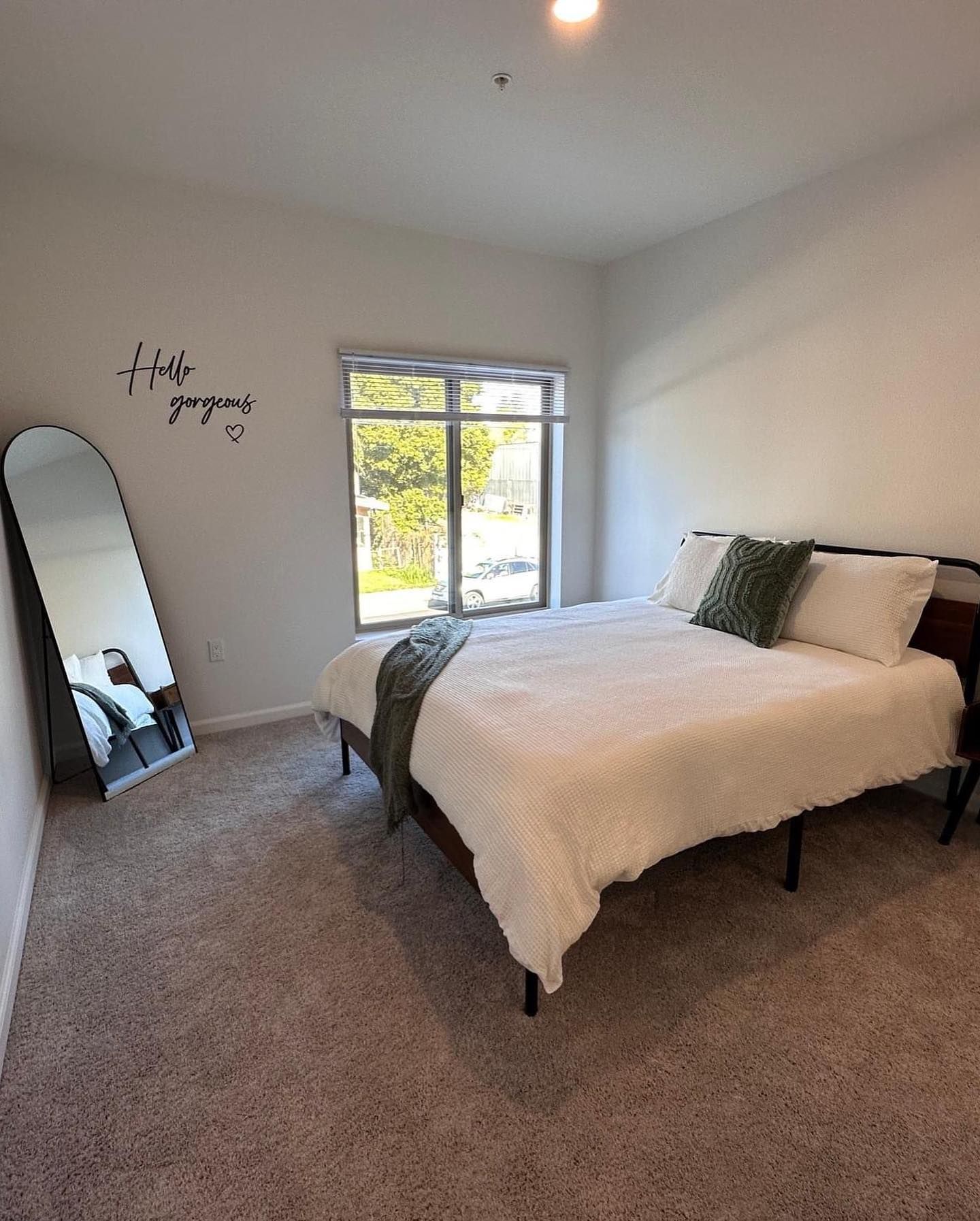 Bright bedroom with a bed, full-length mirror, window, and beige carpet.