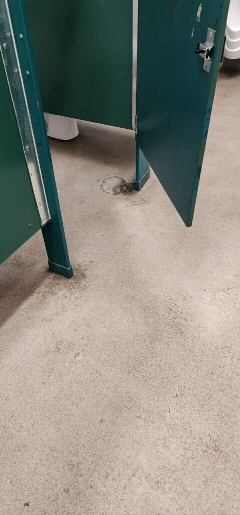 Open green restroom stall doors over a beige tile floor, with a dirty floor area beneath.