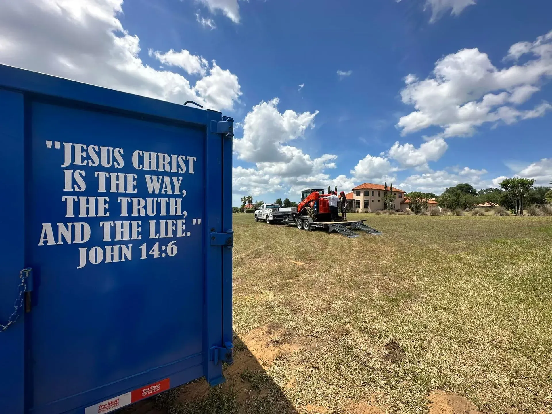 Blue container with religious quote, construction equipment on field with house in background.