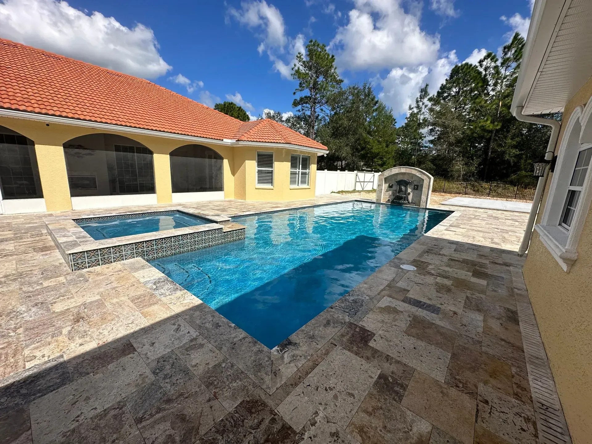 Pool and patio with yellow house and red tile roof under blue sky.