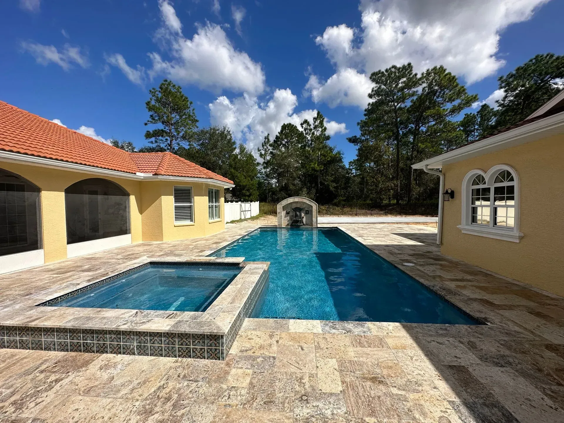 Pool with spa, surrounded by yellow house, red tile roof, and trees under a blue sky.