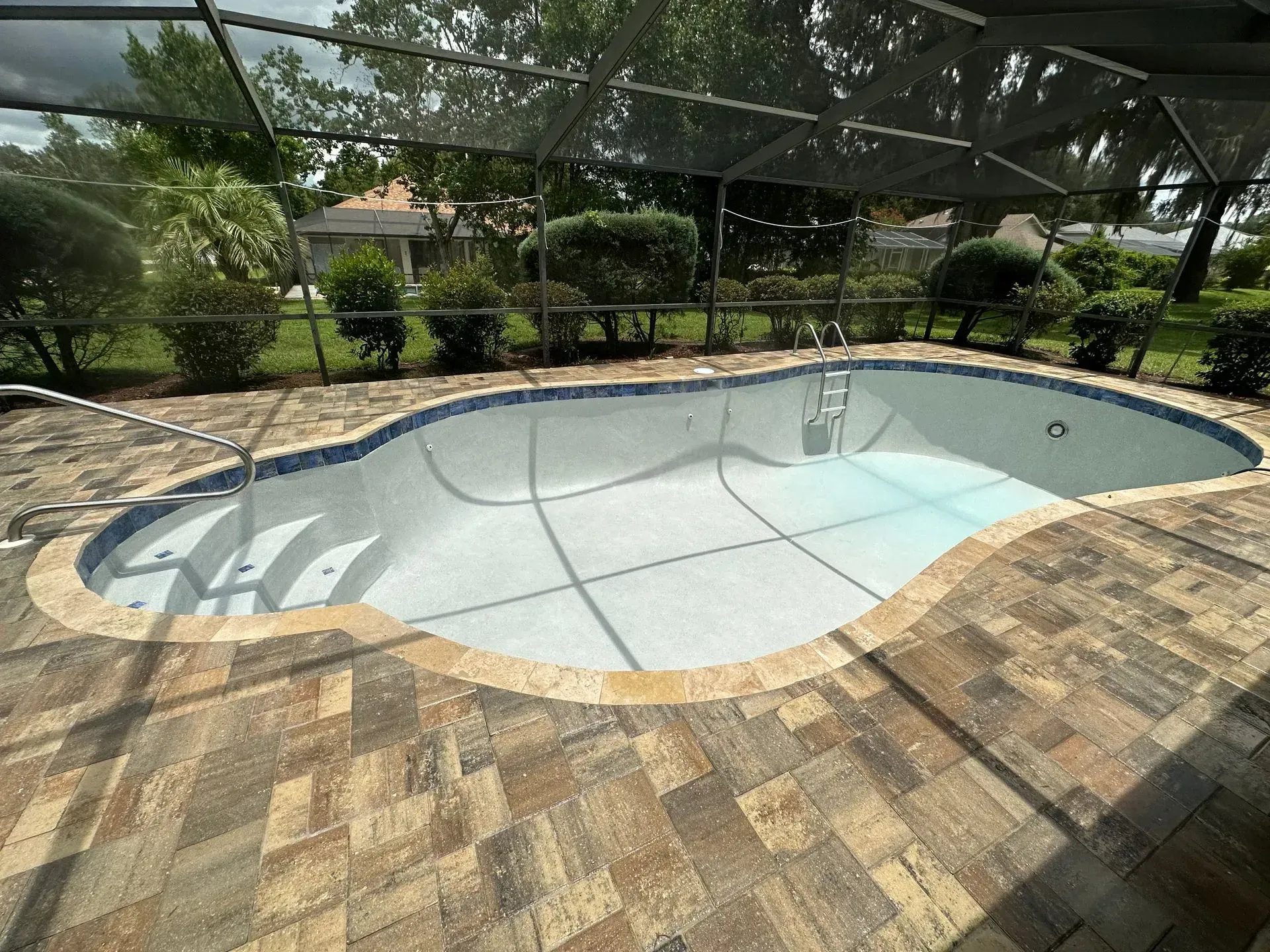 Empty swimming pool with brick patio, surrounded by bushes under a screened enclosure.
