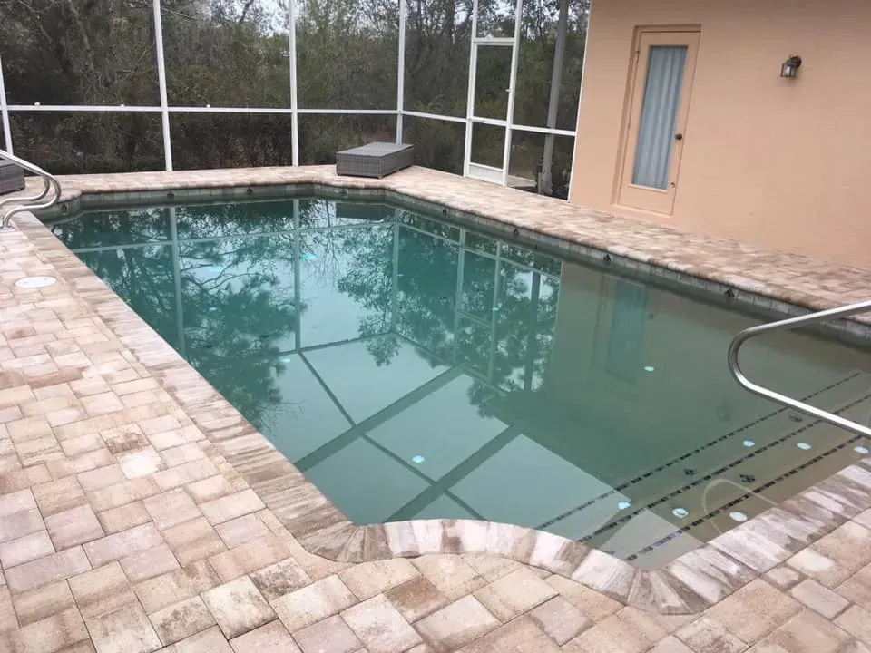 Swimming pool surrounded by brick paving and screened enclosure. Exterior view of water with stairs.