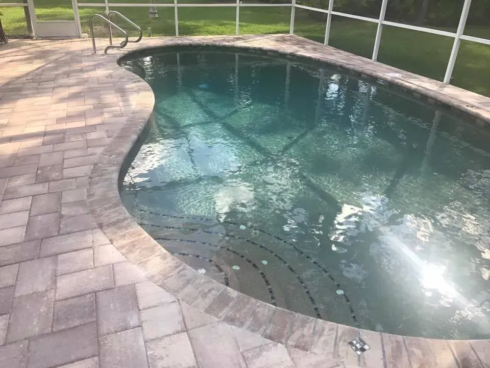 Pool with gray brick surround, turquoise water. Steps lead into the pool. Screened enclosure in background.