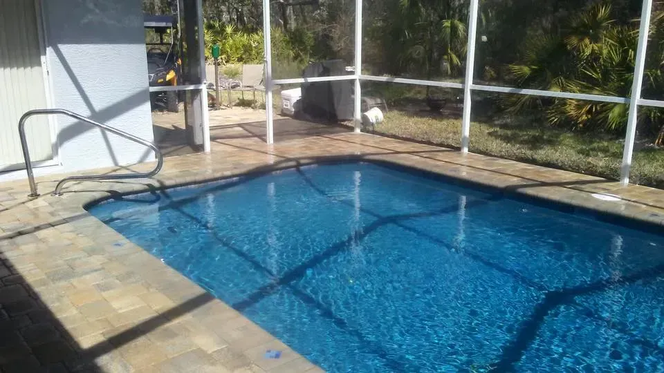 Blue rectangular pool surrounded by a beige stone patio and screened enclosure. Handrail on the side.