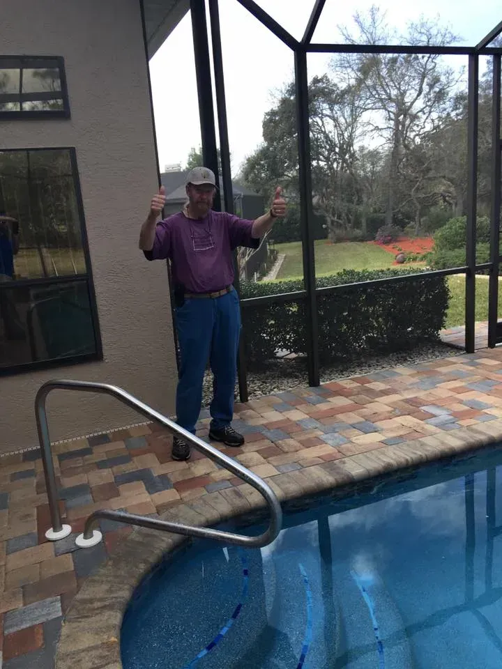 Man standing by pool, giving thumbs-up gestures, with a screened enclosure and backyard visible in the background.