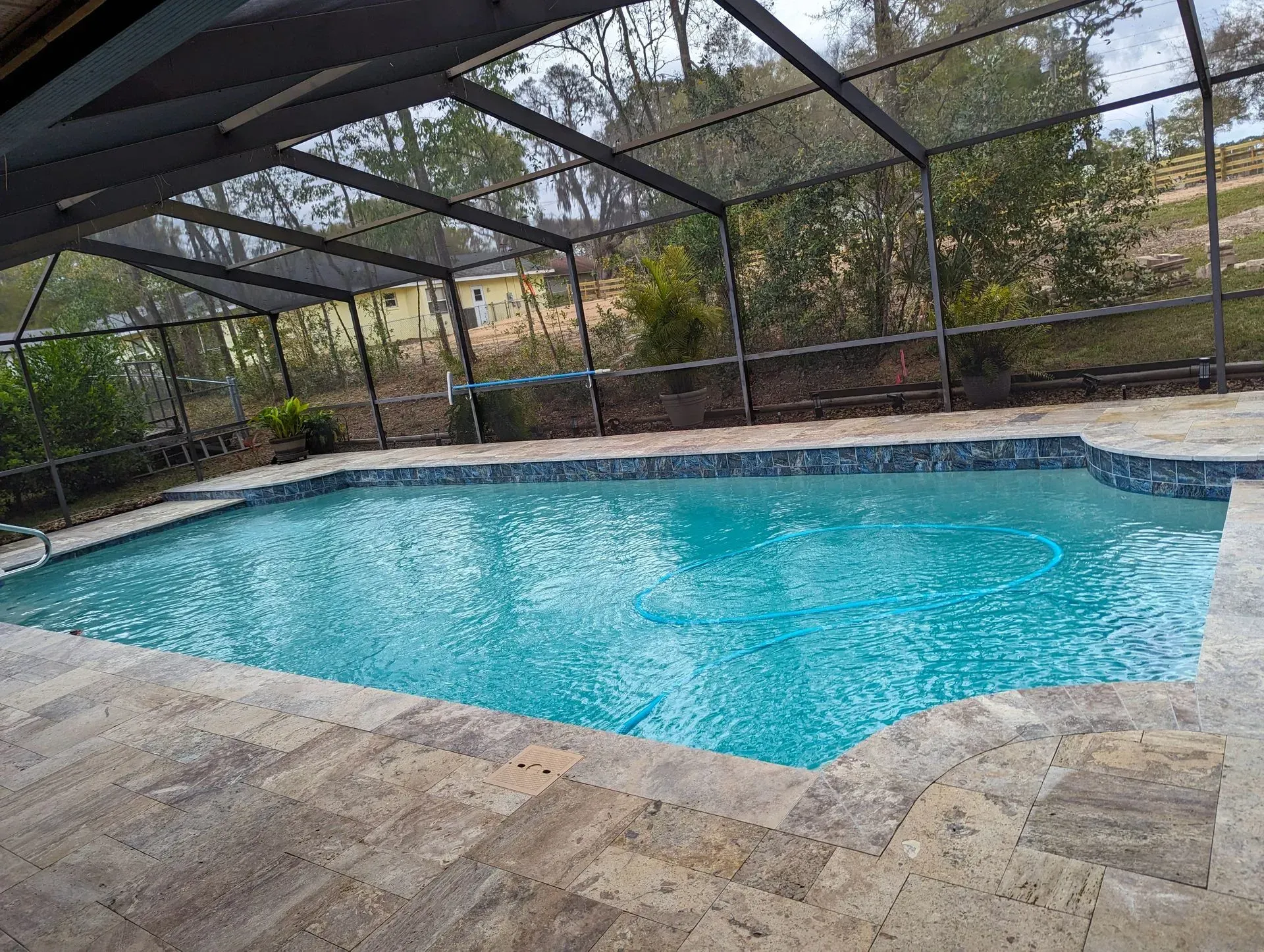 Pool enclosed by a screen. Blue water, stone patio. Green trees in the background.