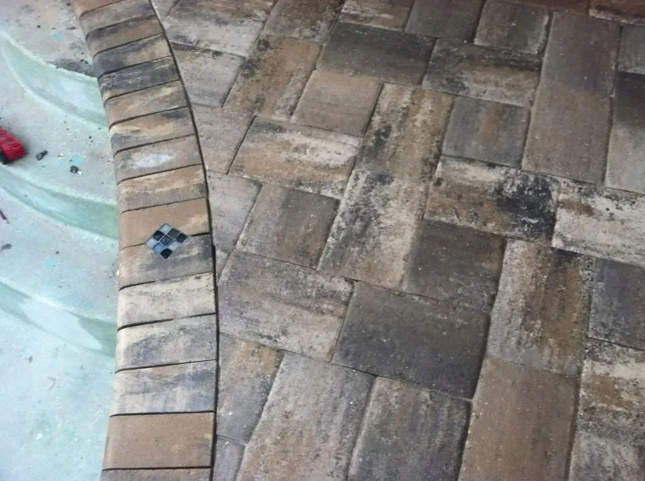 Brick pavers surround a pool, with a curved edge and a random pattern. Brown, beige, and gray tones are visible.