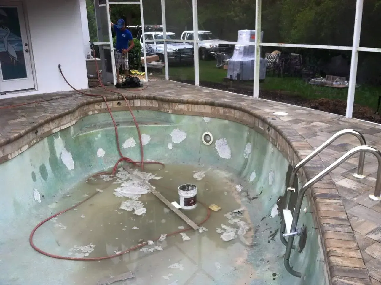 Empty swimming pool with peeling paint, surrounded by brick and a screened enclosure. A person works nearby.