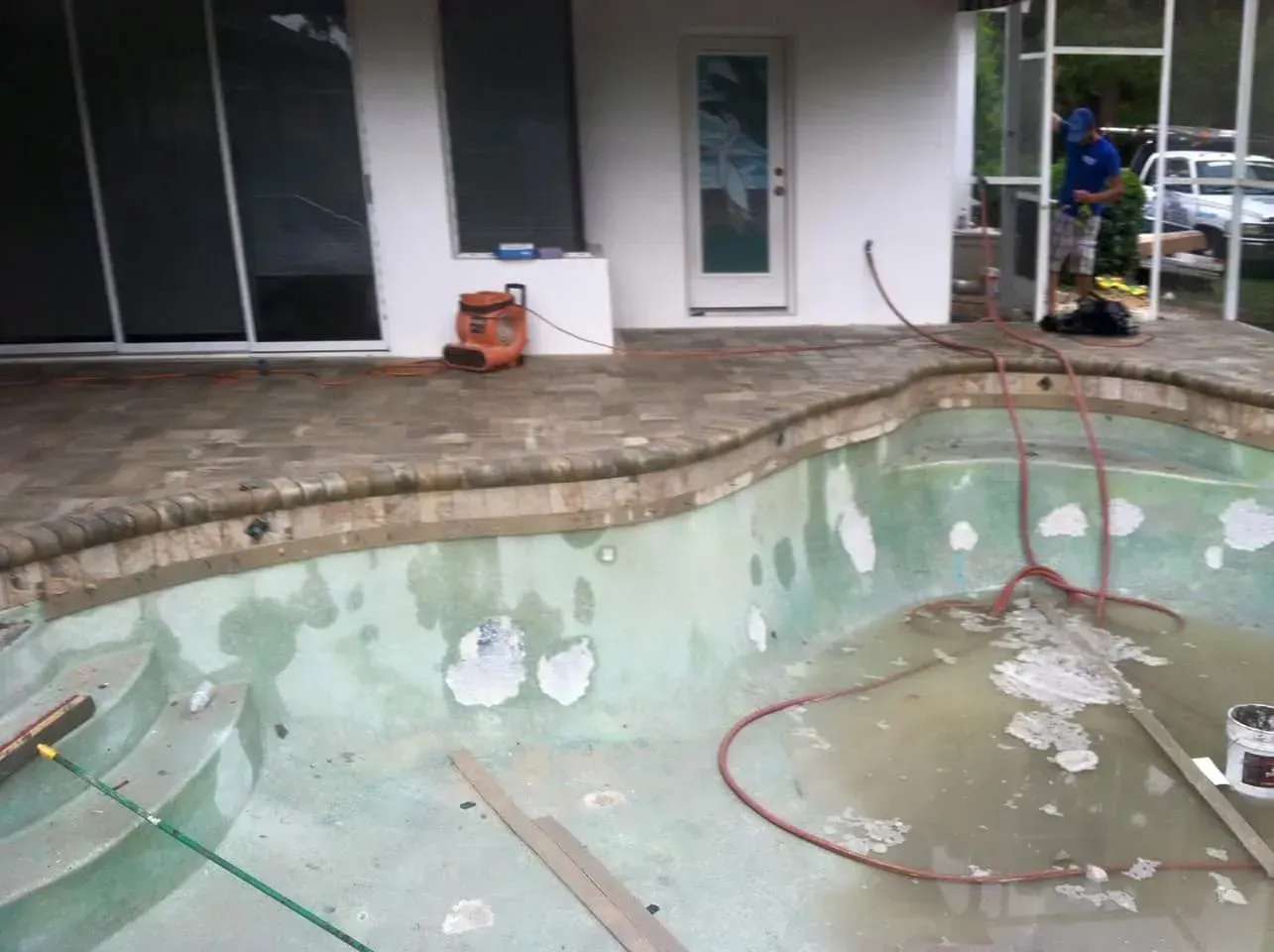Pool undergoing renovation with peeling surface. Worker visible. Outdoor setting.
