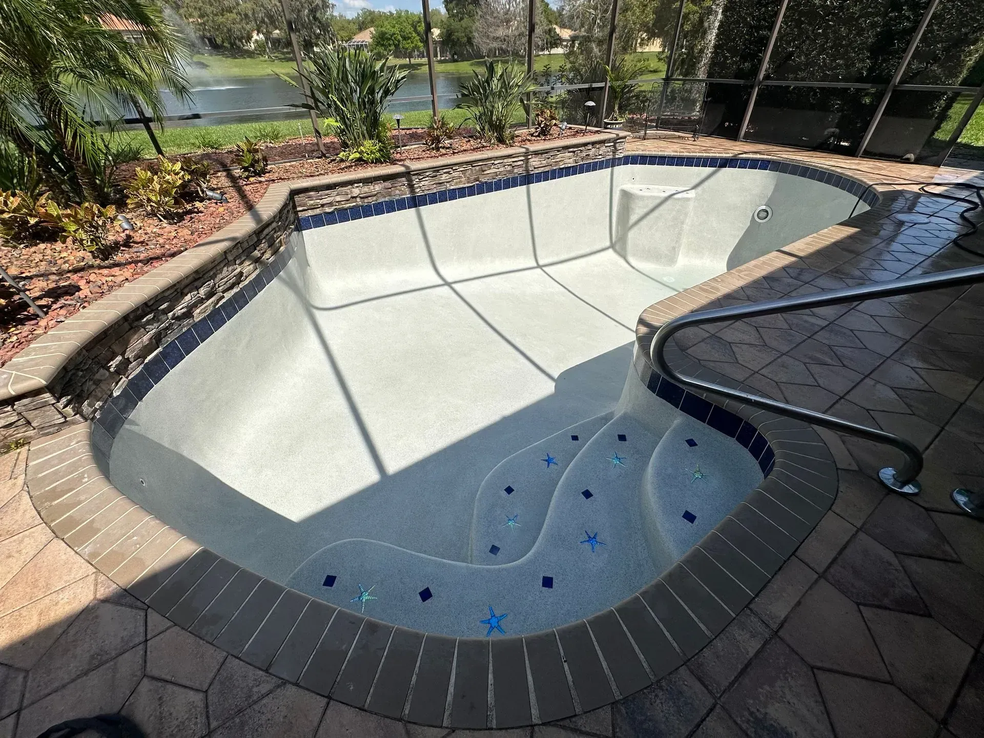 Empty, in-ground pool with stone coping and mosaic tile detail, steps with a stainless steel handrail, and a view of a lake.