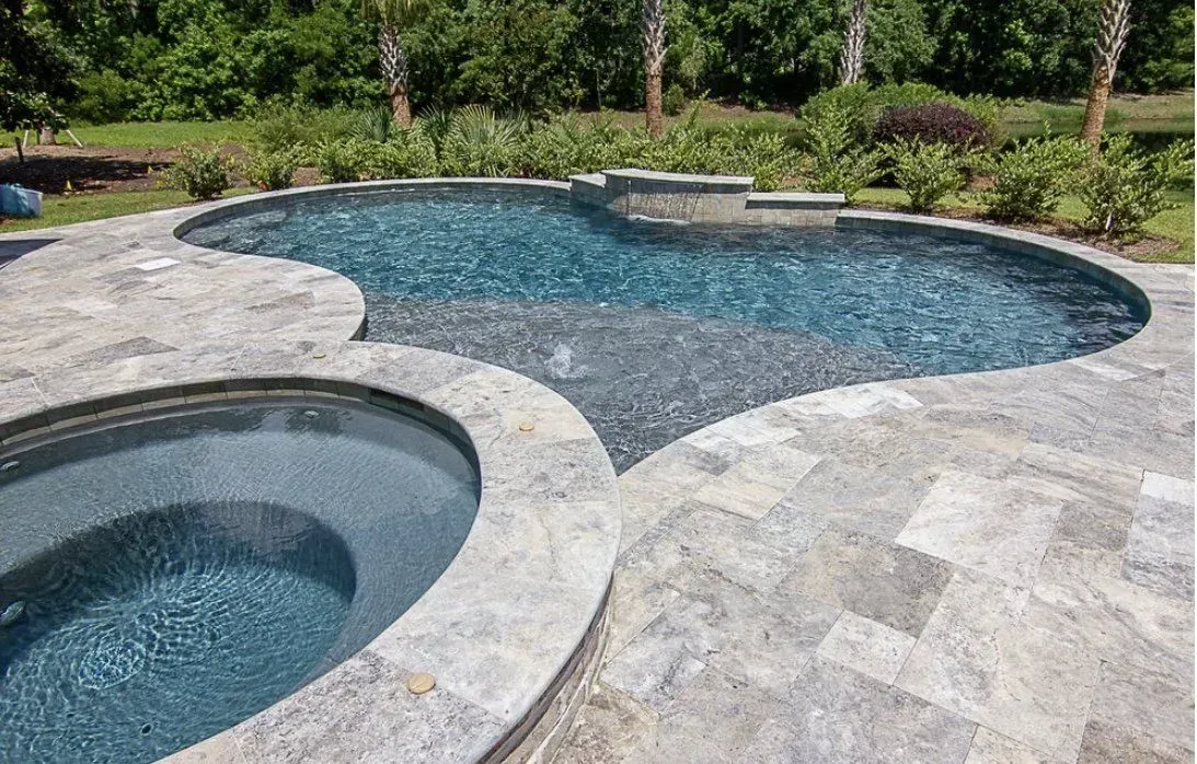 Pool and spa with gray stone patio, surrounded by greenery. Blue water, sunny day.