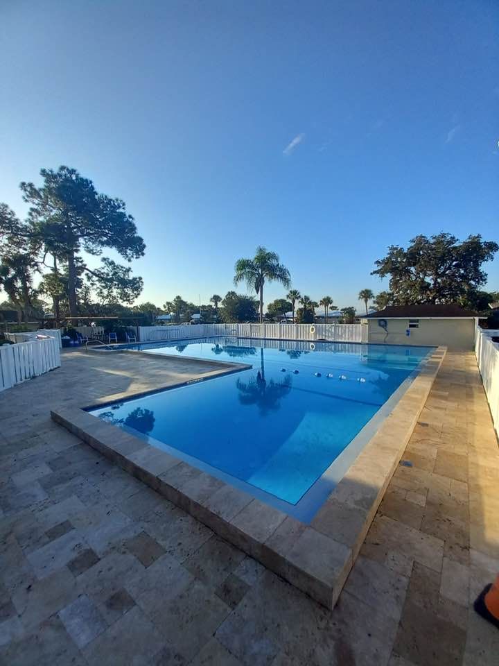 A large, blue swimming pool outdoors on a sunny day. Beige tiled deck surrounds the pool.