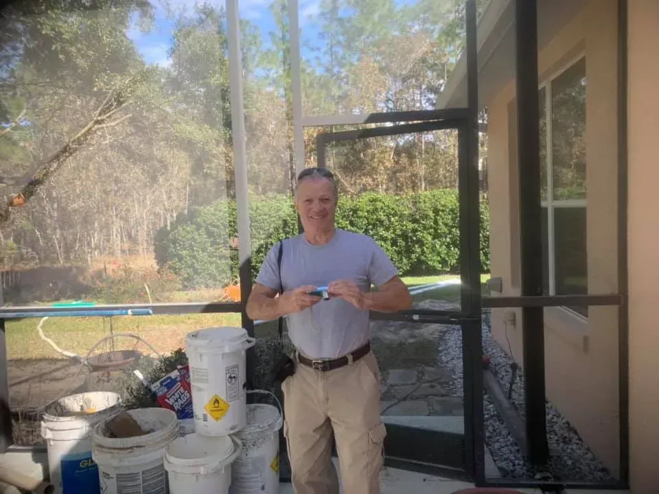 Man holding small object near screened porch. Buckets and debris on ground, trees in background.