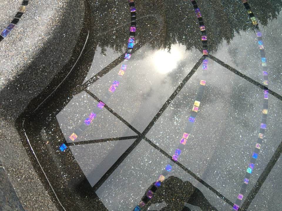 Reflective water surface with mosaic tiles, reflecting trees and sky, on speckled dark stone.