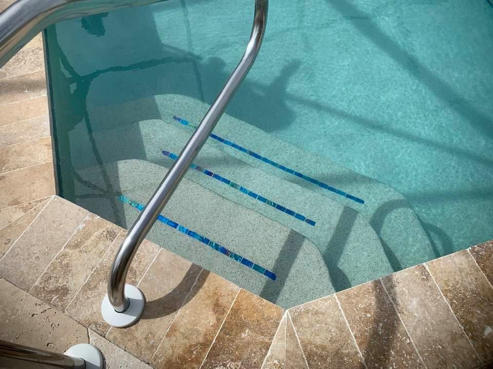 Pool steps with a metal handrail. Beige tile surrounds pool with clear water. Blue tile strips on steps.