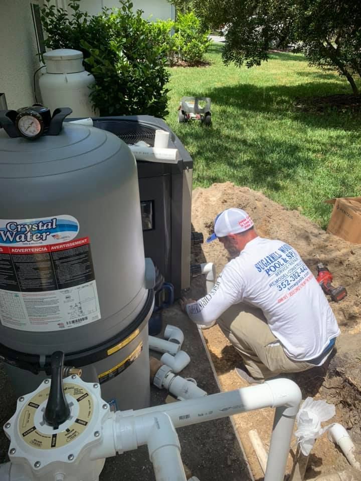 Pool technician working on plumbing near a gray pool filter. He is wearing a white shirt and hat.