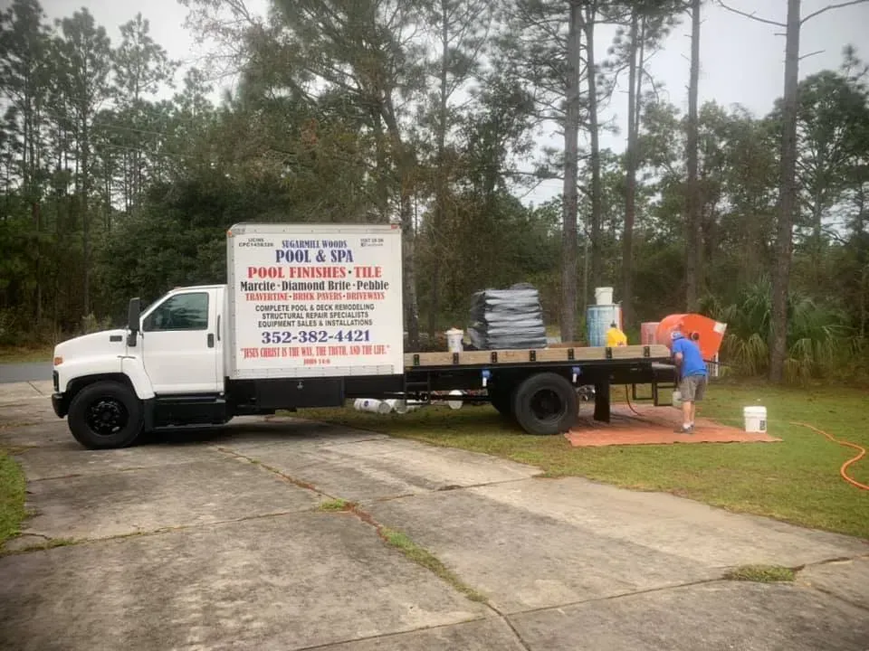 White truck with a flatbed; a person is working on the bed in a yard with trees.