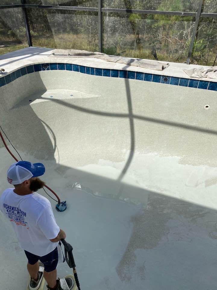 Man applying resurfacing material to the interior of an empty swimming pool with a sprayer; a screened enclosure is in the background.