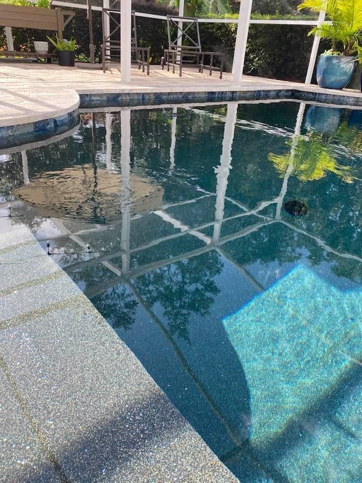 Swimming pool with blue tiles, reflecting a white screened enclosure and greenery. Stone border.