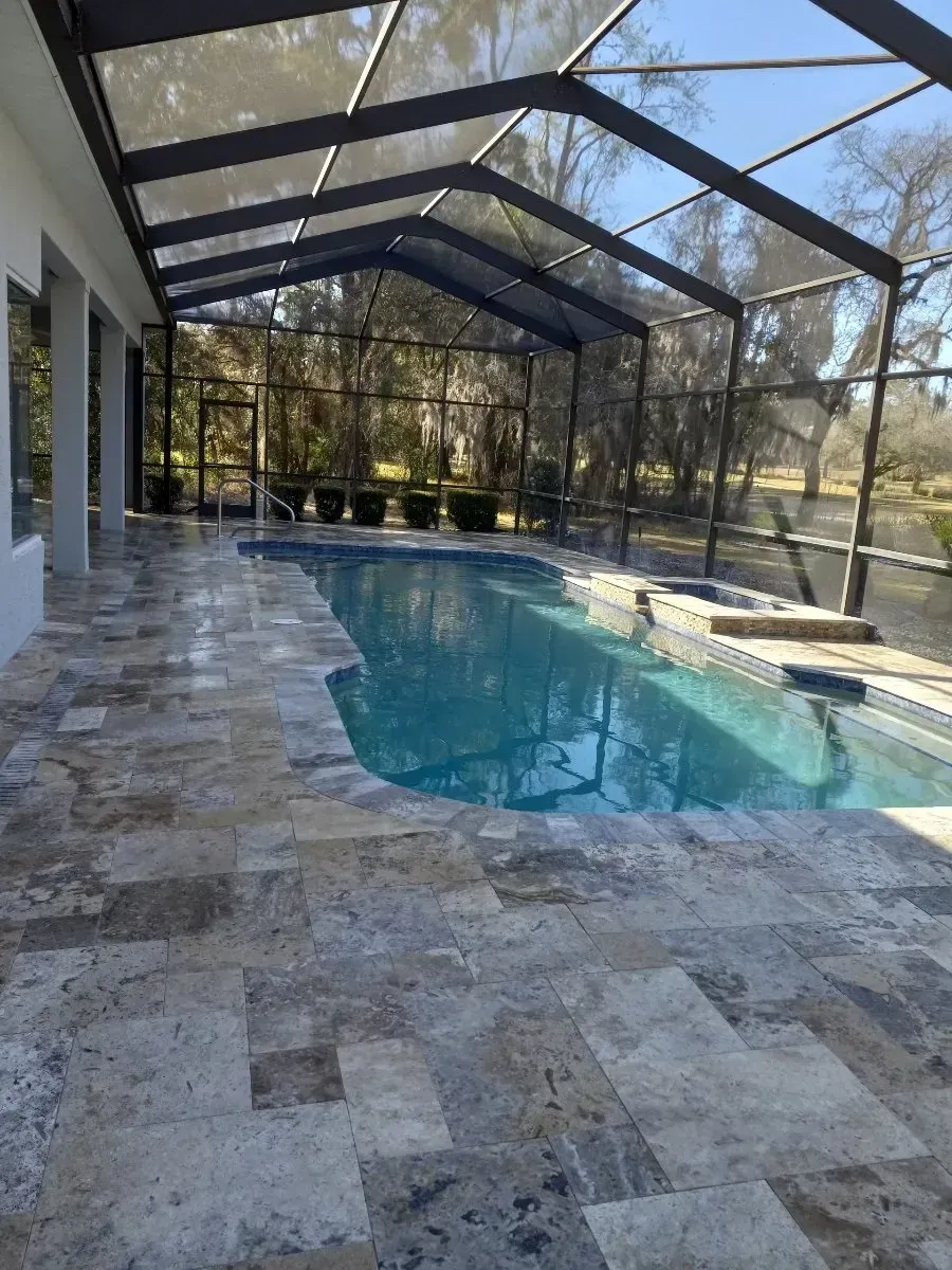 Pool area with a screened enclosure. Stone patio surrounds a rectangular pool, surrounded by trees.