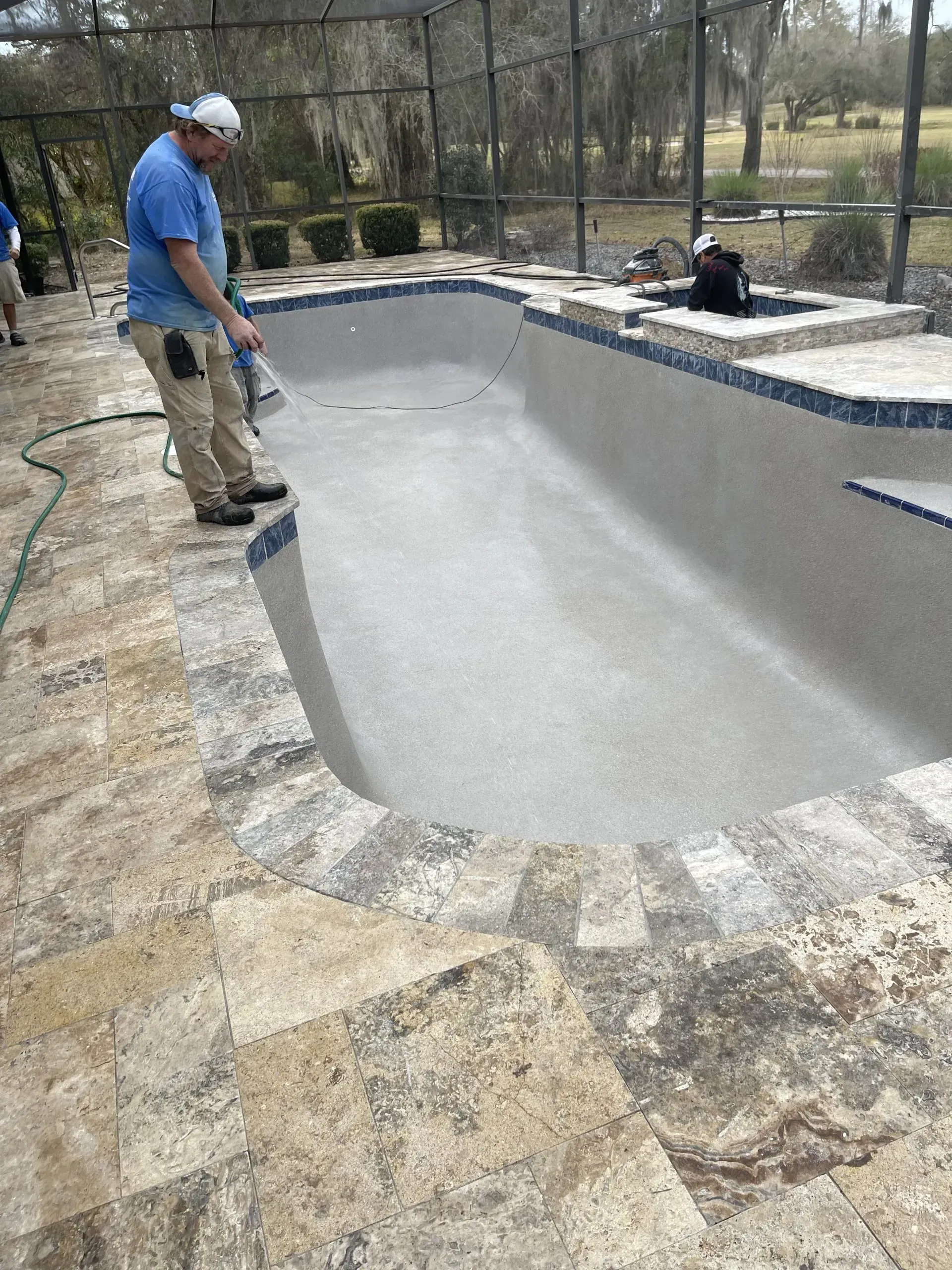 Man spraying a newly plastered swimming pool, surrounded by stone patio, trees, and fence.