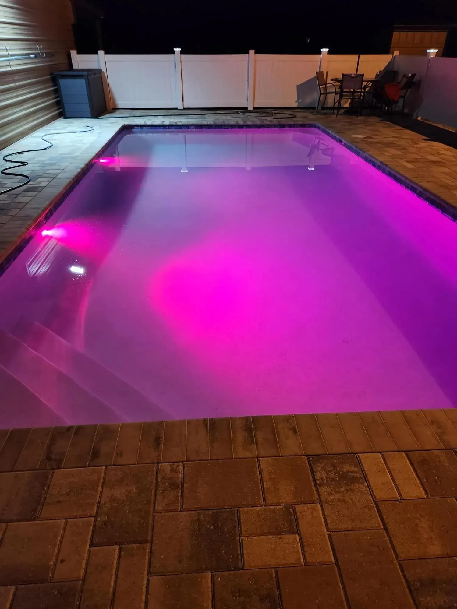 Lit purple pool at night, surrounded by brick patio and white fence.