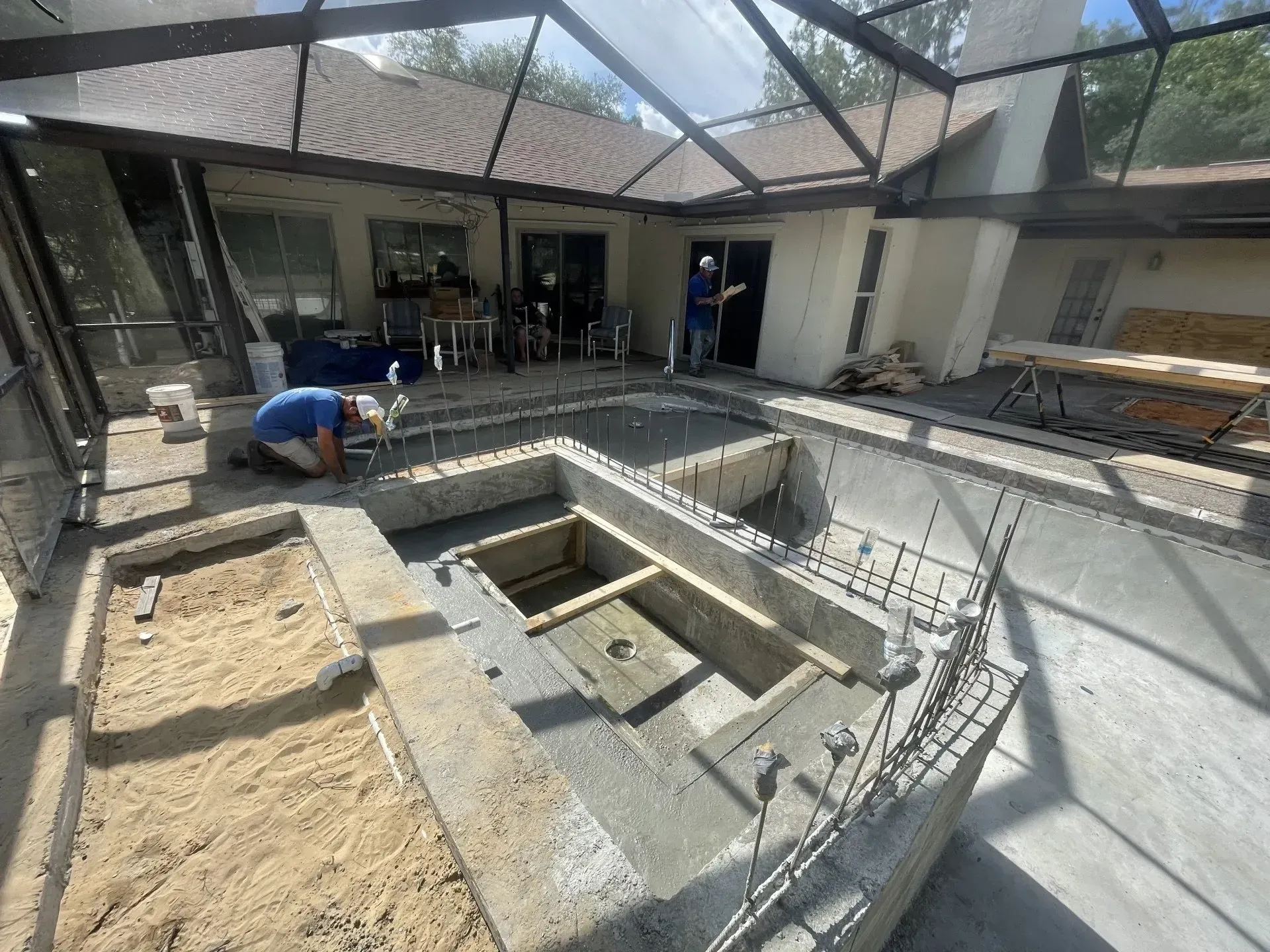 Pool renovation in progress: workers, concrete structure, and patio area under a screened enclosure.