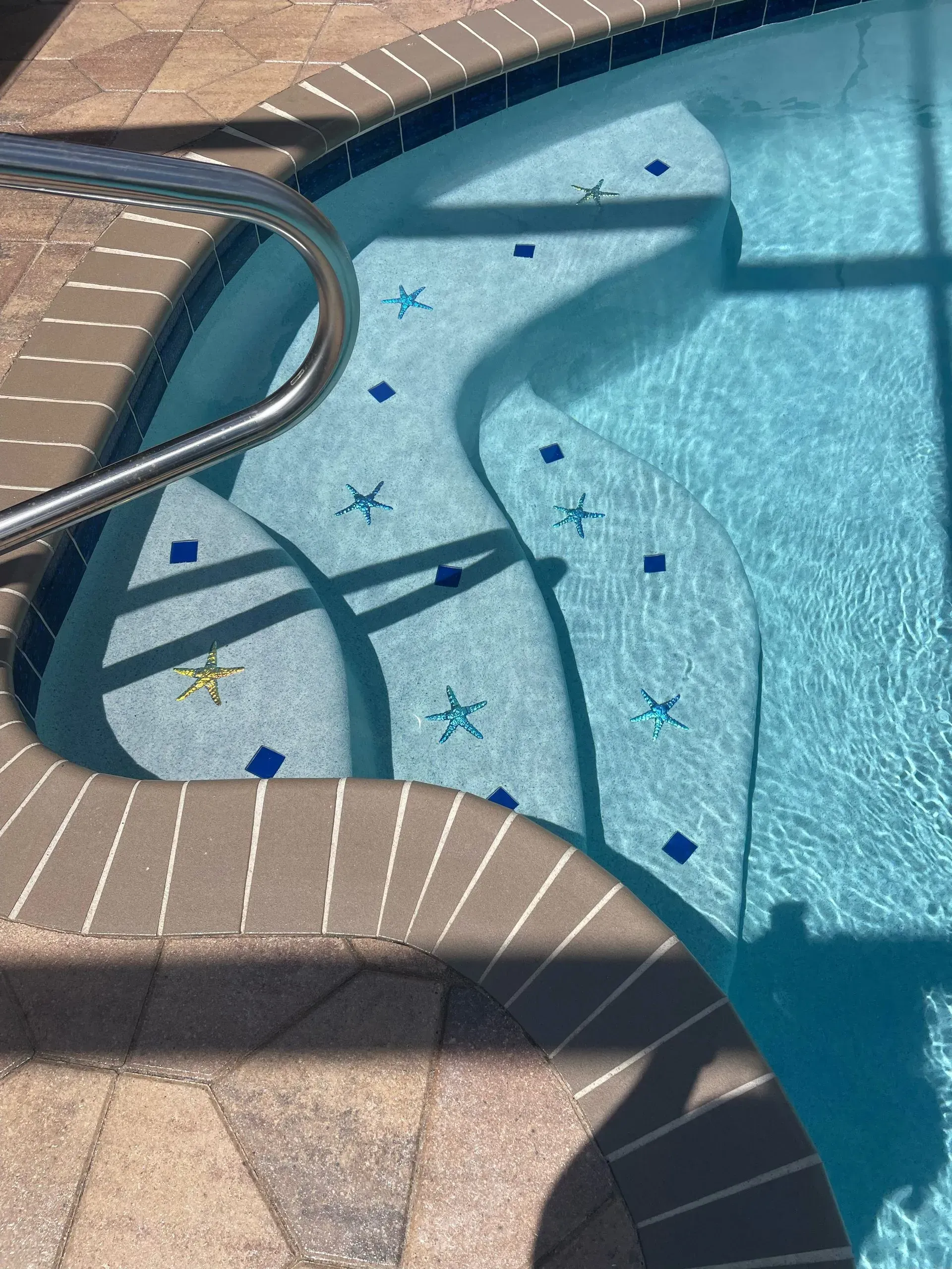 Pool steps with blue water, decorated with starfish and blue tiles, a metal handrail, and brick edging.