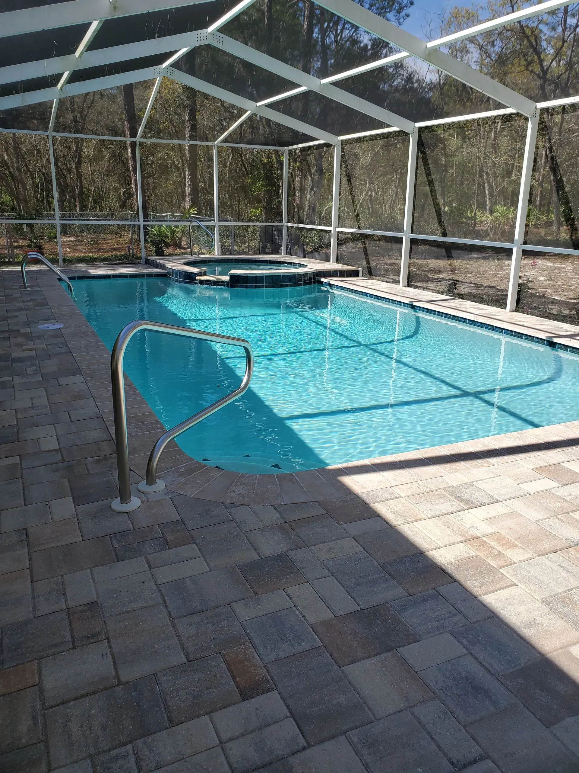 Pool enclosed by a screen. Blue water, brick patio, spa, and trees in background.