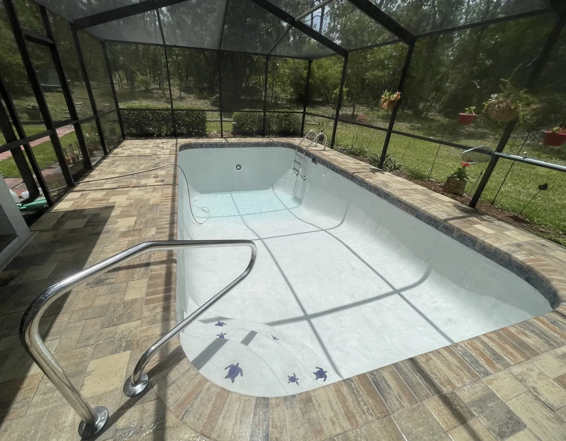 An empty rectangular pool within a screened enclosure, surrounded by tiled patio, with a metal handrail.