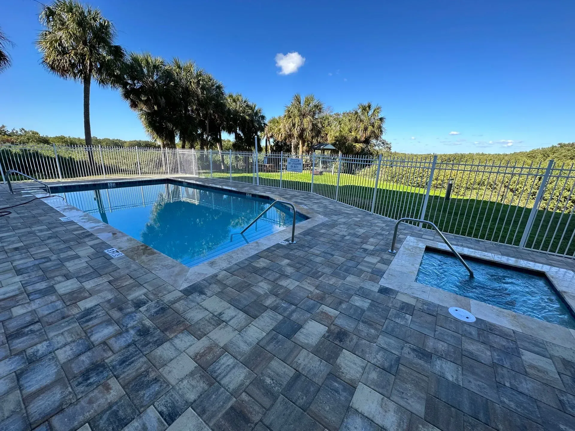 Pool and jacuzzi surrounded by patterned brick, fence, and trees under a blue sky.