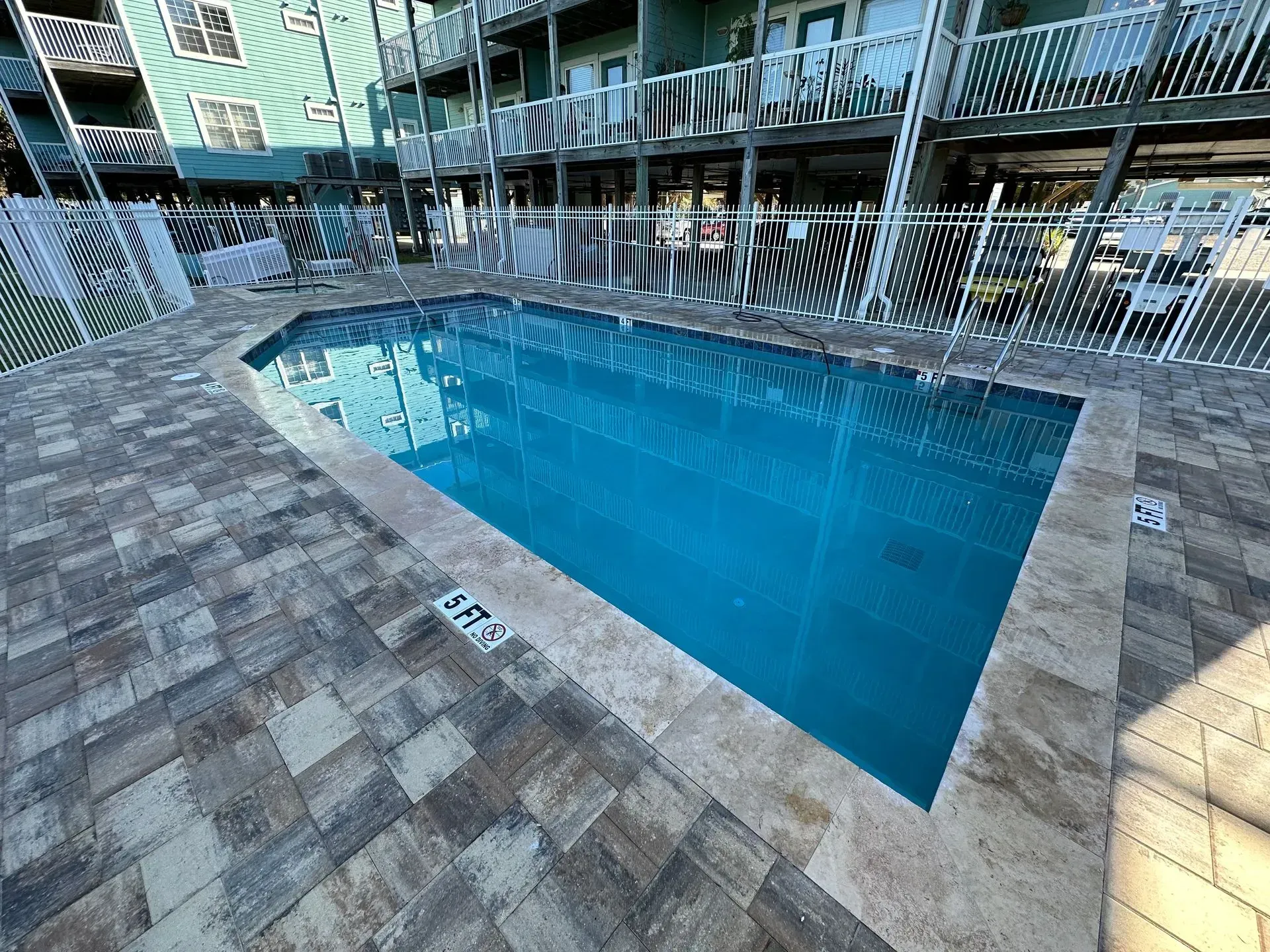 A rectangular swimming pool surrounded by paved tiles; buildings and fencing in background.