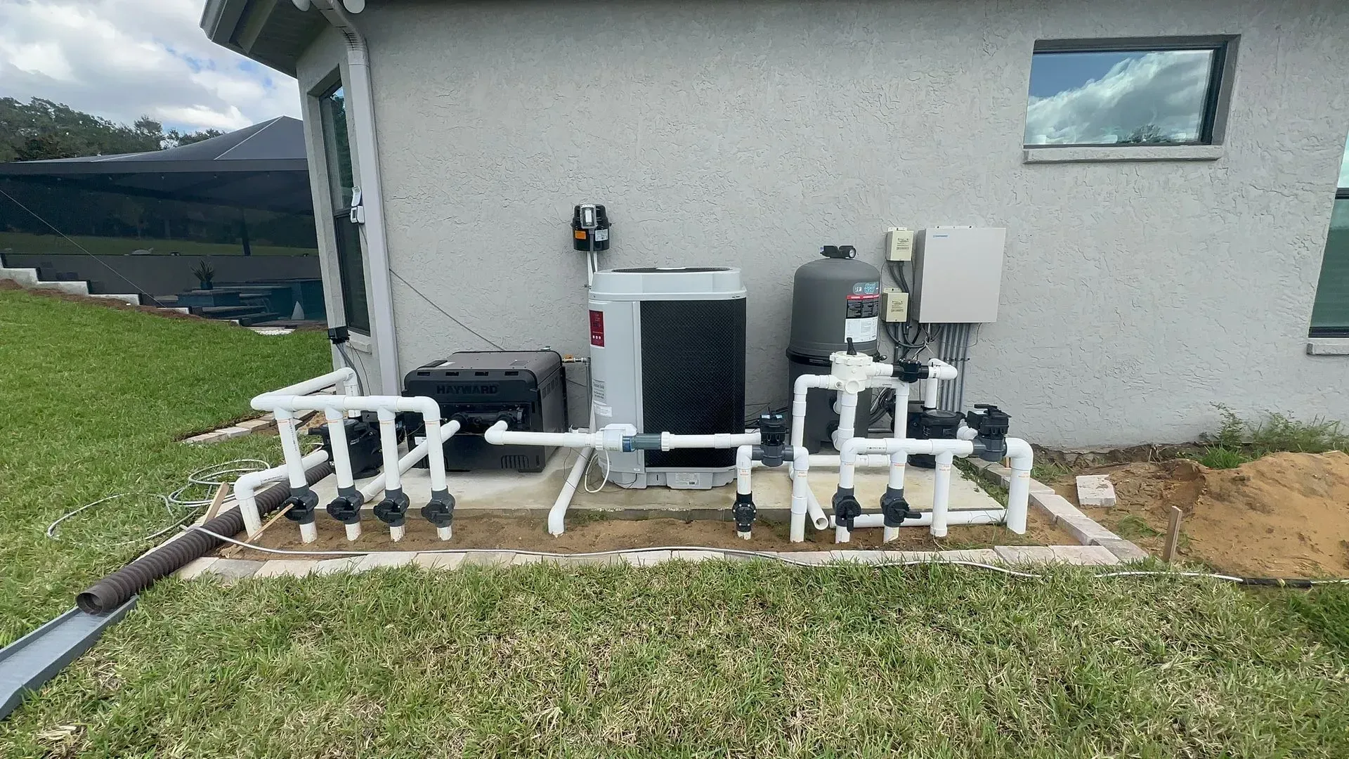 Pool equipment installation against a light-colored building, including pumps, filter, and heater.