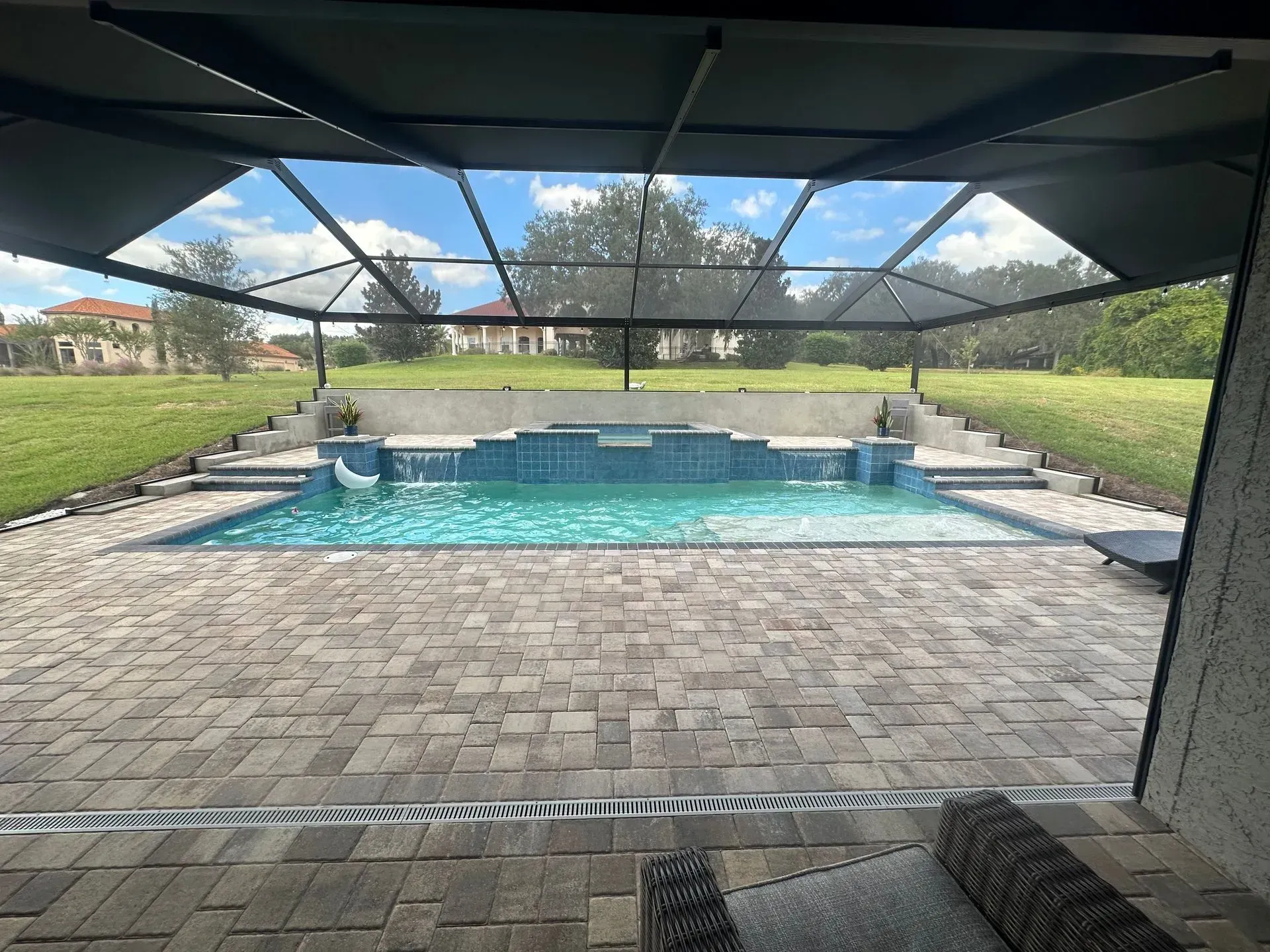 Pool with blue tiled waterfall, screened enclosure, and stone patio overlooking a grassy backyard.